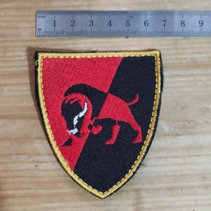 Peut inclure: Un écusson brodé en forme de bouclier avec un motif divisé en diagonale rouge et noir et une silhouette de bison au centre. L'écusson est bordé d'un fil jaune et mesure environ 6,5 cm de largeur, comme indiqué par la règle visible.