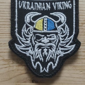 Peut inclure: Un écusson brodé noir en forme de bouclier avec le texte UKRAINIAN VIKING en blanc. Le motif représente un guerrier barbu stylisé portant un casque à cornes avec des segments jaune et bleu, le tout souligné par un fil blanc.