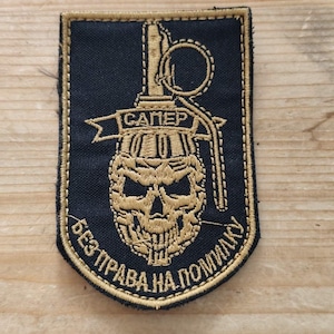 May include: A rectangular black embroidered patch with gold stitching featuring a skull integrated into a hand grenade design. The text "САПЕР" appears on a banner, and "БЕЗ ПРАВА НА ПОМИЛКУ" is arched below. A metal ruler shows the patch is 5 cm wide.