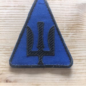 Peut inclure: Un écusson brodé triangulaire avec un fond en tissu bleu vif et une bordure noire cousue. Au centre de l'écusson se trouve un symbole de trident brodé en noir, un emblème à trois branches avec une pointe centrale verticale en forme de lance.