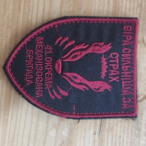 May include: A shield-shaped fabric patch with a black background and dark red embroidered borders and text. The patch features a central stylized animal head graphic and Cyrillic text that reads 41 ОКРЕМА МЕХАНІЗОВАНА БРИГАДА and БІРАСИЛЬНІША ЗА СТРАХ.
