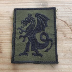 Peut inclure: Un écusson rectangulaire vert olive orné d'un dragon noir brodé de profil. Le dragon est représenté debout sur ses pattes arrière, les ailes déployées et la queue enroulée. L'écusson présente une bordure noire épaisse sur tout le pourtour.
