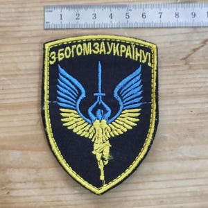 Peut inclure: Un écusson brodé en forme de bouclier avec un fond noir et une bordure jaune. Il représente une figure centrale aux ailes bleues et jaunes tenant une épée, avec le texte "З БОГОМ ЗА УКРАЇНУ!" au-dessus. Une règle montre une largeur de 9 cm.