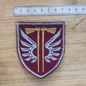Op de afbeelding: Een schildvormige geborduurde patch met een bordeauxrode achtergrond, twee gouden bijlen en lichtblauwe vleugels. Een metalen liniaal erboven toont een breedte van ongeveer 6,5 cm. De patch heeft een lichtblauwe gestikte rand.