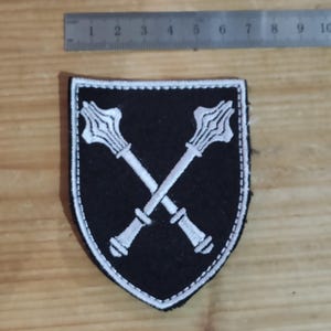 May include: A black shield-shaped patch featuring white embroidered stitching of two crossed maces. A metal ruler is positioned above the patch, showing it measures approximately 2.5 inches in width and 3.5 inches in height.