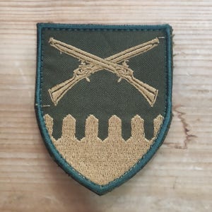 May include: A shield-shaped embroidered patch featuring a dark olive green background with a dark green border. The design includes two crossed antique muskets in tan thread above a stylised tan crenellated wall pattern at the bottom edge.