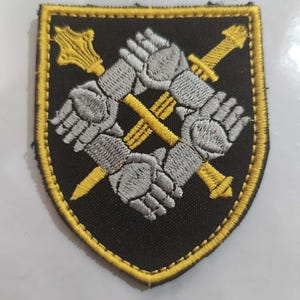Peut inclure: Un écusson brodé en forme de bouclier avec un fond noir et une bordure jaune. Le motif présente quatre gantelets blindés gris disposés en carré, enserrant deux masses d'armes jaunes croisées au centre de l'écusson.