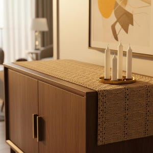 Puede incluir: Un aparador de madera con herrajes dorados está cubierto con un camino de mesa tejido. Cuatro velas blancas están en un soporte dorado. Una obra de arte abstracta cuelga de la pared. Una silla y una mesa son visibles.