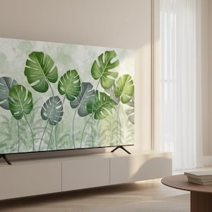 Puede incluir: Un televisor de pantalla plana grande muestra una impresión botánica de hojas de monstera verdes y grises. El televisor está montado en un mueble beige claro en un salón moderno. También se ven una mesa de centro redonda y un sofá.
