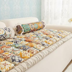 Könnte beinhalten: Ein weißes Sofa mit einem farbenfrohen, gemusterten Kissen und Kissenrollen. Das Kissen hat ein Patchwork-Design mit verschiedenen geometrischen und floralen Mustern in Orange, Gelb, Blau und Braun. Eine blaue Vase mit weißen Blumen steht daneben.