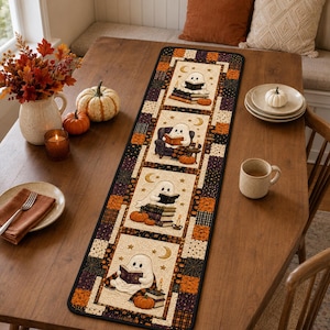 May include: A rectangular table runner featuring a patchwork border in orange, black, and cream with spiderweb patterns. The center panels display illustrations of white ghosts reading books, surrounded by crescent moons, stars, and orange pumpkins.