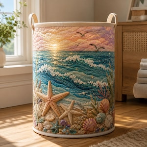 May include: A cylindrical laundry hamper featuring a quilted fabric exterior with a coastal scene. The design depicts a sandy beach with starfish and seashells in the foreground, ocean waves, and a sunset sky with birds flying overhead.