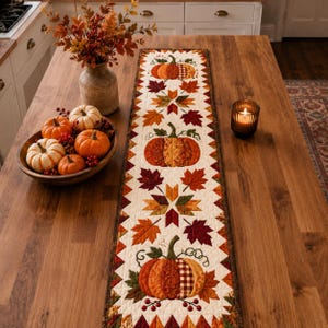 May include: A rectangular quilted table runner featuring a repeating pattern of orange pumpkins and multicolored autumn leaves on a cream background, bordered by a dark brown edge with a triangular geometric design along the perimeter.