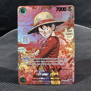 Może przedstawiać: Karta kolekcjonerska One Piece z Monkey D. Luffy. Karta ma czerwone i złote tło foliowe z ilustracją Luffy'ego w słomkowym kapeluszu. Tekst: "RED HAWK!!!!" i "Monkey D. Luffy".