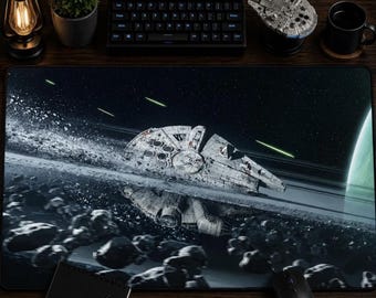 May include: A black rectangular mouse pad featuring a detailed illustration of the Millennium Falcon spaceship in space. The design includes a planet, asteroids, and laser fire. A black keyboard, mouse, and other desk accessories are also visible.