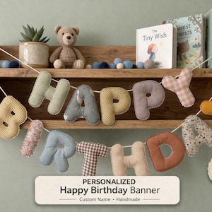Puede incluir: Un cartel personalizado de "Feliz Cumpleaños" con letras de tela en varios patrones y colores. El cartel cuelga frente a una estantería de madera con libros, un oso de peluche y una planta en maceta. El cartel está diseñado con tonos neutros.