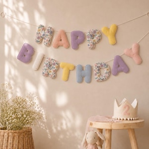 Pode incluir: Uma guirlanda de tecido com a inscrição HAPPY BIRTHDAY em letras pastel e florais suspensa por um cordão. Inclui uma coroa de tecido bege com o número 2, um coelho de pelúcia com vestido rosa e uma cesta de vime com flores secas.