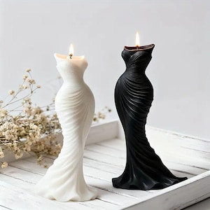 May include: Two dress-shaped candles, one white and one black, on a white tray. The candles are lit, with flames at the top. The dresses have a draped design. The background is a soft white.