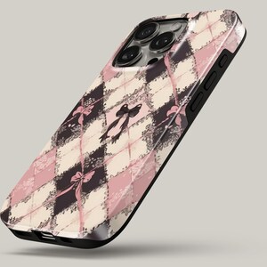 May include: A smartphone case featuring a repeating argyle pattern in shades of dusty pink, cream, and dark charcoal. The design includes delicate black lace-like floral accents and small pink ribbon bows at the intersections of the diamond shapes.