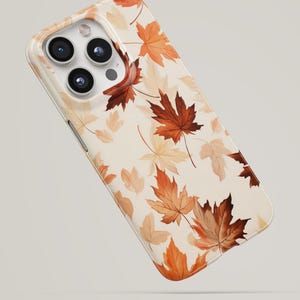 Puede incluir: Una funda para smartphone con fondo color crema y un patrón repetitivo de hojas de arce otoñales en tonos naranja, óxido y marrón claro. La funda tiene un recorte para un sistema de cámara de triple lente y un flash en la esquina superior.