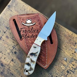 Könnte beinhalten: Ein feststehendes Messer mit silberner Stahlklinge, Messingbacke und cremefarbenem Griff mit dunkelbraunen Fleckenmustern. Das Messer liegt auf einer hellbraunen Lederscheide mit Korbgeflecht-Textur und dem Schriftzug Frontier Blades.