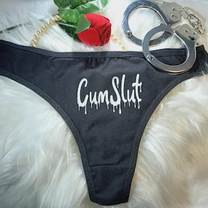 May include: Black thong underwear with the text "Cum Slut" in white dripping font. Silver handcuffs are visible in the background, along with a red rose and a string of pearls.