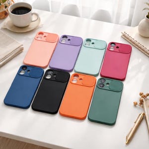 Puede incluir: Una colección de fundas de teléfono coloridas en varios tonos, incluyendo rosa, morado, azul, negro, naranja y verde. Cada funda cuenta con una cubierta para la lente de la cámara. Las fundas están dispuestas sobre una superficie blanca.