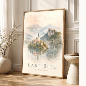 Può includere: Una stampa artistica da parete rettangolare verticale incorniciata con un'illustrazione ad acquerello del Lago di Bled in Slovenia. L'opera raffigura una chiesa su una piccola isola, montagne e un castello su una scogliera, con il testo LAKE BLED SLOVENIA.