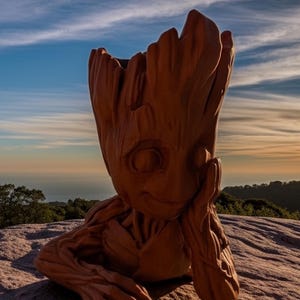 May include: A brown, wooden sculpture of Groot from Guardians of the Galaxy. The sculpture is in a seated position, with its head resting on its hand. The background features a scenic view of a mountain range and a colorful sky.