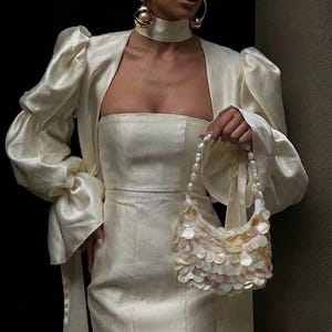 May include: Cream-coloured outfit comprising a strapless mini dress, a matching jacket with puffed sleeves, and a choker. A small, decorative handbag with a pearl strap and shell-like embellishments is also visible. Gold hoop earrings add a touch of elegance.