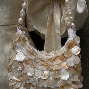 May include: A cream-colored handbag with a rounded shape, covered in iridescent, circular shell pieces. The bag has a pearl beaded handle. The background is a soft, neutral color.