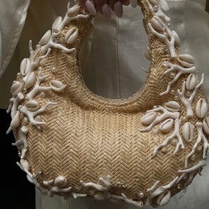 Peut inclure: Un sac à main beige tissé avec une poignée incurvée, orné de coquillages blancs, d'ornements en forme de corail et de petites perles. Le sac présente un motif en chevrons.