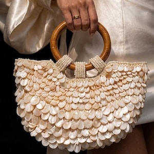 May include: A cream-coloured handbag with a wooden handle and a body covered in small, shell-like embellishments. The handle is a light brown, circular shape. The bag is held by a person wearing a cream-coloured dress.