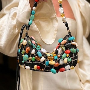 May include: A black handbag decorated with a variety of colourful gemstones, including turquoise, red, yellow, and white stones. The bag has a beaded strap and a black cord tassel. The bag is approximately 25 cm wide.