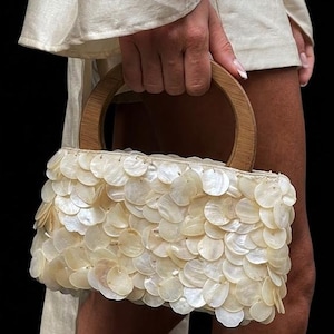 Pode incluir: Uma bolsa creme com uma alça de madeira e um corpo coberto por peças iridescentes sobrepostas, semelhantes a conchas. A bolsa é segurada contra um fundo escuro, mostrando sua textura e design.