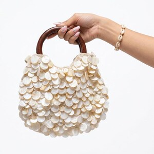 Pode incluir: Uma bolsa creme com formato arredondado, coberta por pequenas decorações redondas em forma de concha. Possui uma alça de madeira marrom escura, semicircular. Uma pulseira de concha combinando também é visível.