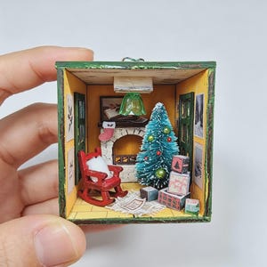 May include: A miniature diorama box featuring a cosy room with a red rocking chair, a small green Christmas tree with ornaments, a fireplace with a hanging stocking, a green ceiling lamp, and several wrapped gift boxes on a white rug.