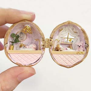 May include: A miniature diorama inside a hinged walnut shell featuring two rooms with pink quilted walls. The left side has a gold lamp, a small plant, and a bed. The right side includes a window with white curtains, a bed, and a small pink bear.