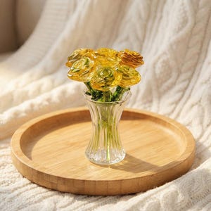 May include: A small glass vase holds a bouquet of yellow glass roses. The vase sits on a round wooden tray, which rests on a white knitted blanket. The roses have green stems and are a mix of yellow and amber.