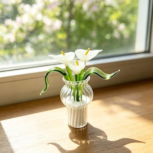 May include: A clear glass vase with vertical ribbing contains three white calla lily flowers made of glass. Each flower features a bright yellow spadix and a long, curved green stem. The vase has a rounded body and a flared rim, holding the flowers upright.