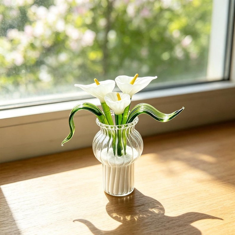 May include: A clear glass vase with vertical ribbing contains three white calla lily flowers made of glass. Each flower features a bright yellow spadix and a long, curved green stem. The vase has a rounded body and a flared rim, holding the flowers upright.