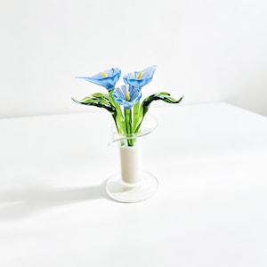 May include: A glass sculpture featuring three light blue calla lily flowers with yellow centers and green stems and leaves, arranged in a clear glass vase with a flared rim and a cylindrical base, set against a plain white background.