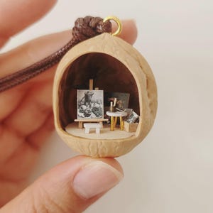 May include: A miniature art studio diorama housed inside a hollowed-out walnut shell pendant. The interior features a tiny easel with a black and white painting, a small white stool, a round table with art supplies, and a box on a light base.