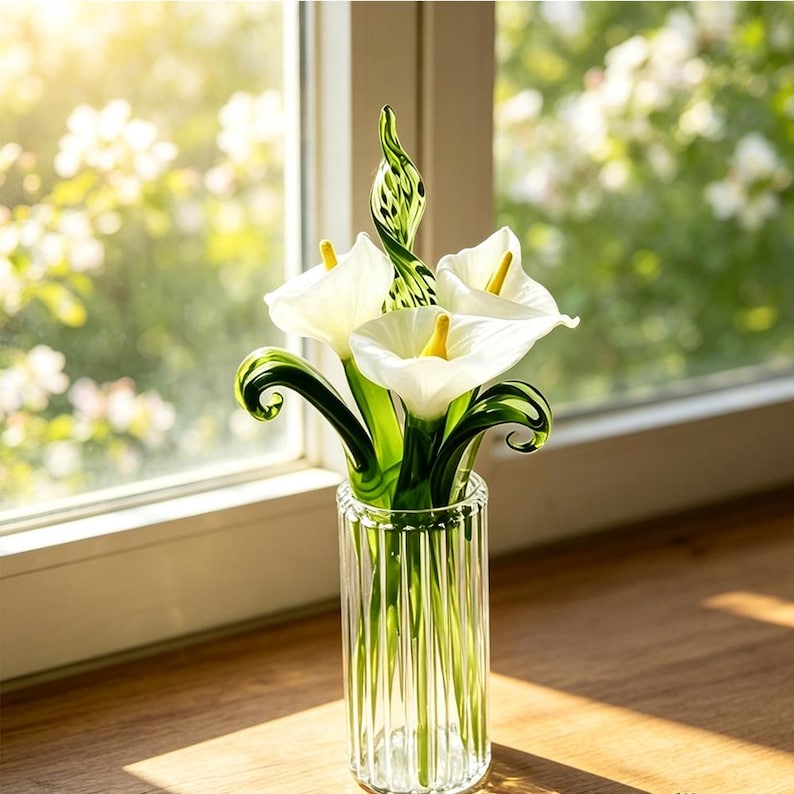 May include: A glass sculpture of three white calla lilies with yellow centers and green stems, featuring swirling green glass leaves and a central spiraled accent, displayed in a clear, vertically ribbed cylindrical glass vase.