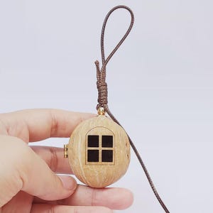 May include: A brown walnut shell pendant featuring a small, arched window cutout with a black interior. The pendant is attached to a dark brown cord with a knotted loop at the top and a small gold-toned metal hinge on the side of the shell.