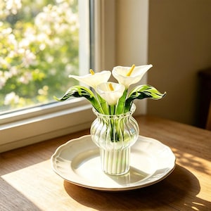 May include: A clear glass vase with vertical ribbing holds three white glass calla lilies with yellow centers and green glass stems and leaves. The vase sits centered on a plain white ceramic plate, showcasing the delicate glass craftsmanship.