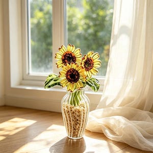 Puede incluir: Un florero de vidrio transparente con estrías verticales que contiene tres girasoles amarillos con centros de color marrón oscuro y hojas verdes. La base del florero está llena de pequeñas piedras claras y los tallos son visibles.