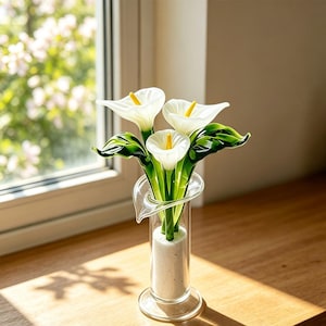 May include: A clear glass vase containing three white glass calla lilies with yellow centers and green glass leaves. The vase has a cylindrical body with a flared rim and is filled with a white granular material at the base to hold the flowers.