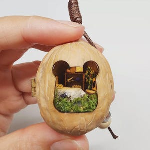 May include: A miniature diorama housed inside a hollowed-out walnut shell, featuring a tiny room with a bed, a wooden shelf, and green foliage. The walnut shell is hinged with a small metal clasp and includes a brown cord for hanging as a pendant.