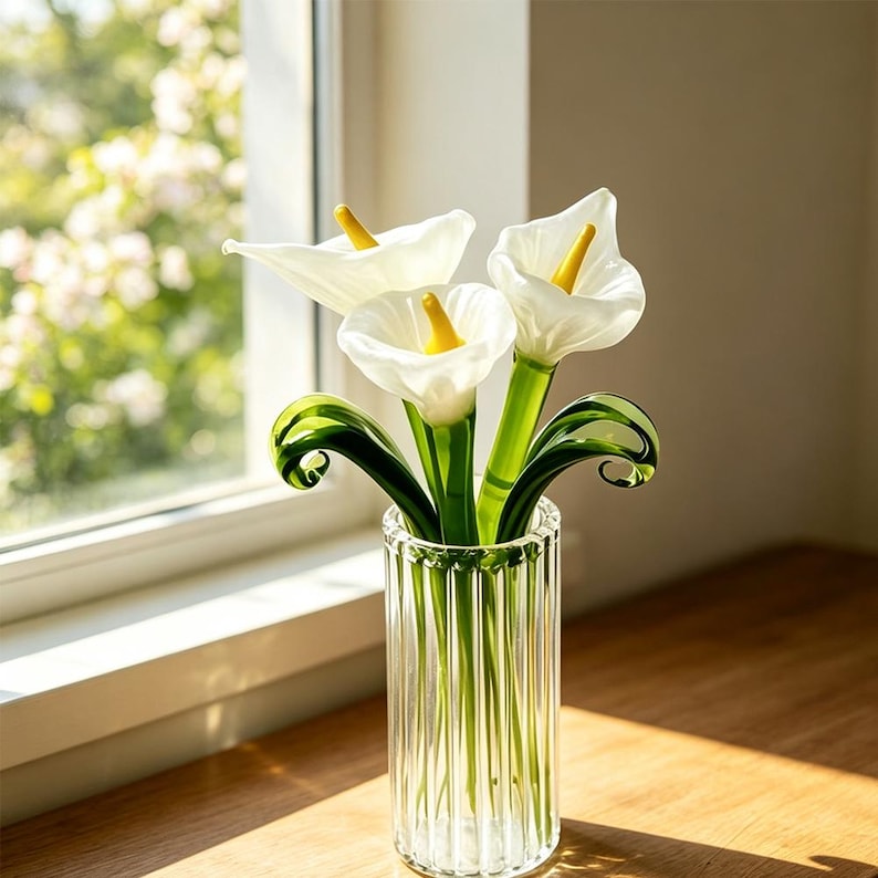 May include: A clear, vertically ribbed glass vase containing three glass calla lilies. Each lily features a smooth, opaque white petal structure with a bright yellow spadix in the center. The stems and curled leaves are made of translucent green glass.
